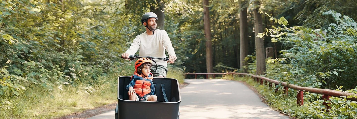 Ein fröhlicher Mann fährt mit einem Kind im Lastenrad einen Waldweg entlang.