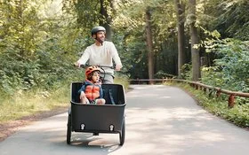 Ein fröhlicher Mann fährt mit einem Kind im Lastenrad einen Waldweg entlang.
