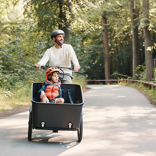 Ein fröhlicher Mann fährt mit einem Kind im Lastenrad einen Waldweg entlang.