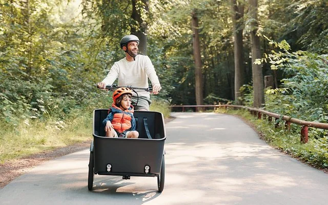 Ein fröhlicher Mann fährt mit einem Kind im Lastenrad einen Waldweg entlang.