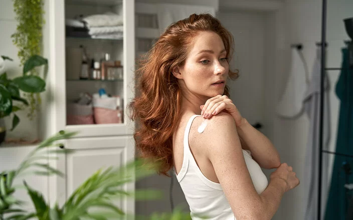 Eine Frau mit langen roten Haaren in einem Badezimmer. Sie cremt sich ihre rechte Schulter mit einer weißen Lotion ein.