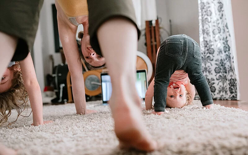 Zwei junge Frauen und ein Kleinkind, die im Wohnzimmer auf einem hellen Teppich Yoga-Übungen machen. Sie stehen auf den Zehenspitzen und beugen den Oberkörper nach unten, die Hände sind am Boden. Die Übung heißt herabschauender Hund.