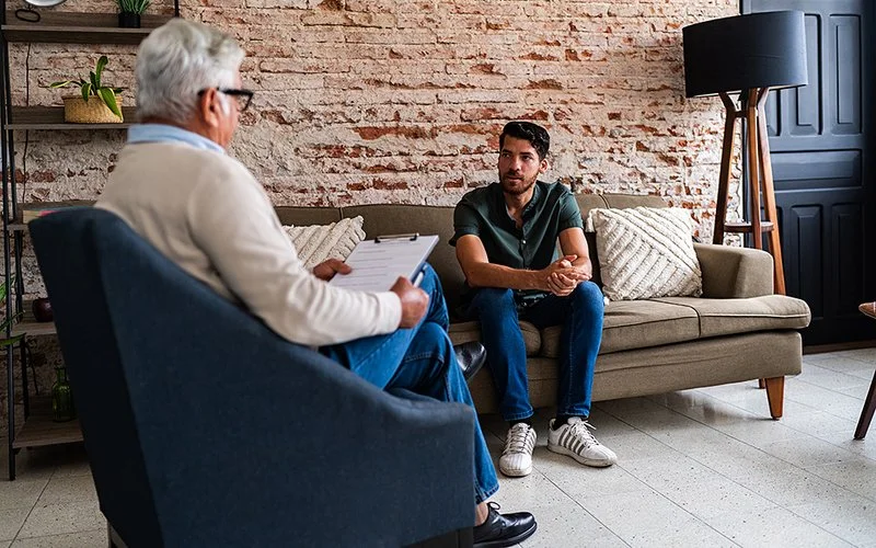 Ein Mann sitzt im Praxisraum eines Therapeuten auf der Couch. Der Therapeut sitzt im gegenüber auf einem Sessel und hält ein Klemmbrett in den Händen.