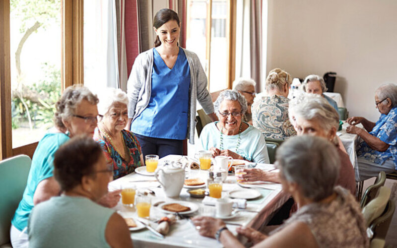 Eine junge Frau steht an einem gedeckten Tisch, an dem sechs ältere pflegebedürftige Frauen sitzen.