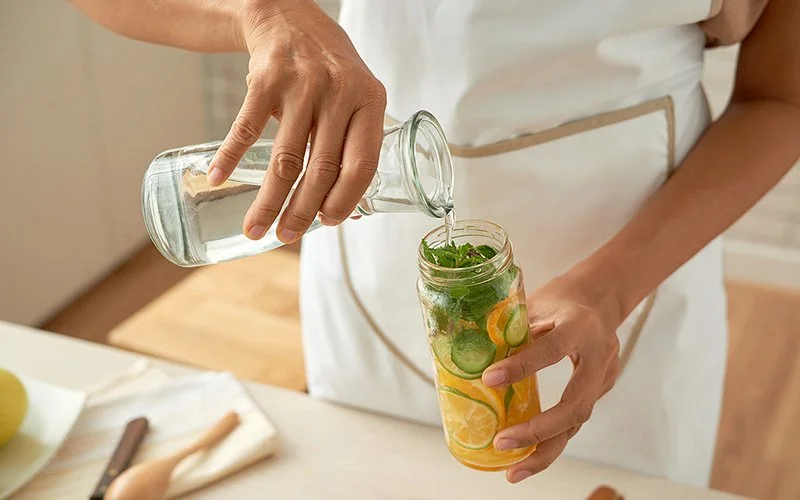Person gießt Wasser aus einer Glaskaraffe in ein Glas mit Zitronen-, Gurkenscheiben und Minzblättern.