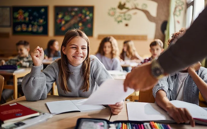 Ein Schulmädchen sitzt an seinem Tisch im Klassenraum. Neben ihr und im Hintergrund sitzen weitere Schüler und Schülerinnen. Eine Person, von der nur der ausgestreckte Arm abgebildet ist, reicht dem Mädchen einen Zettel, den es mit der linken Hand ergreift. Freudestrahlend blickt das Mädchen auf den Zettel.