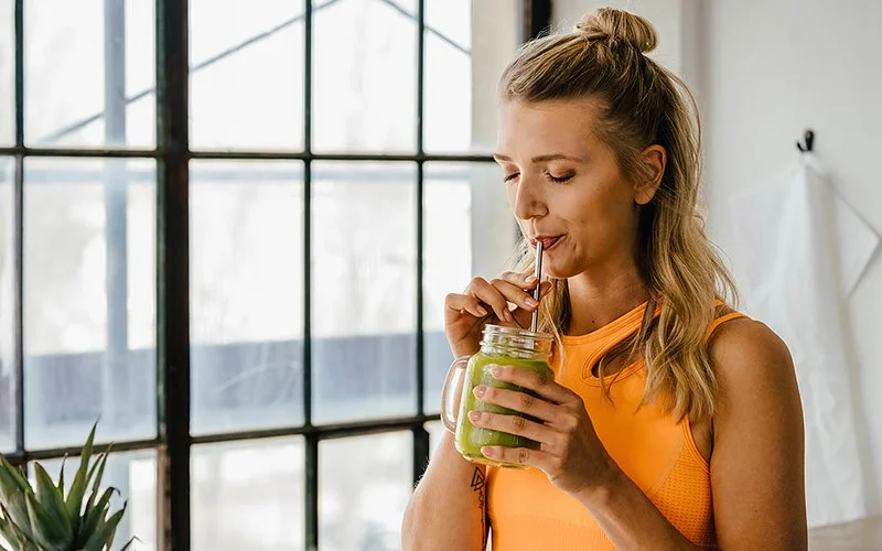 Eine sportlich wirkende junge Frau im orangefarbenen Top steht vor einem Fenster und trinkt einen grünen Smoothie durch einen Strohhalm.