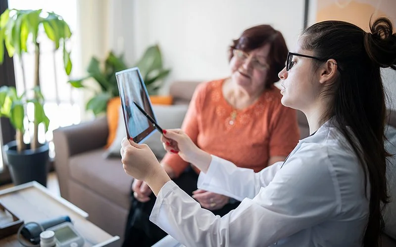Eine Ärztin erläutert einer älteren Patientin das Röntgenbild eines Brustkorbs. Dazu hält sie das Bild ins Gegenlicht und zeigt mit einem Kugelschreiber auf einen Punkt darauf.