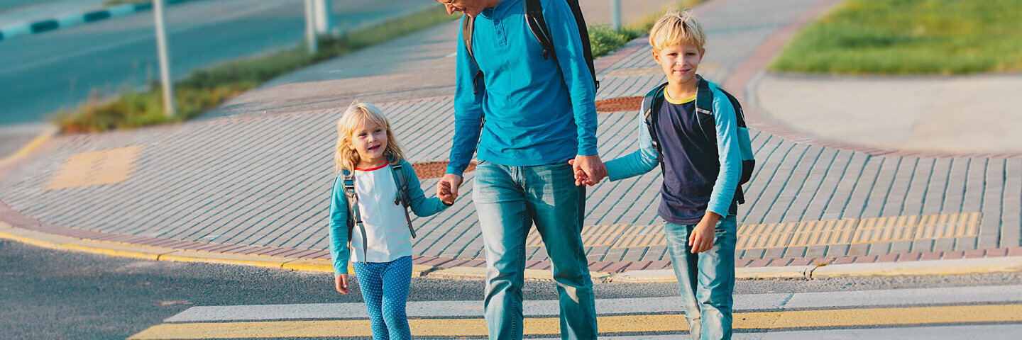 Vater geht mit Sohn und Tochter zur Schule. Die Familie hält sich beim Überqueren der Straße an den Händen.