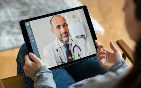 Nahaufnahme eines Tablets, das von einer Hand gehalten wird. Darauf läuft eine Videosprechstunde mit einem Arzt.