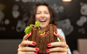 Ein junge Frau in einem Lokal hält lachend ein angebissenes Sandwich in die Kamera.