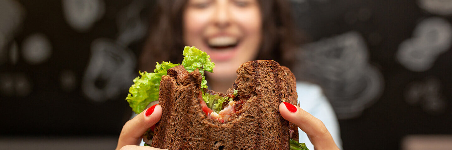 Ein junge Frau in einem Lokal hält lachend ein angebissenes Sandwich in die Kamera.