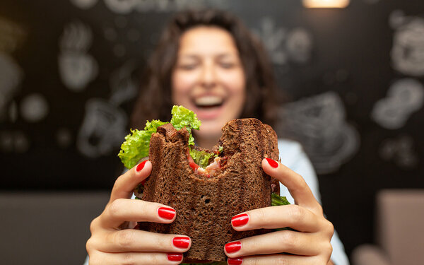 Ein junge Frau in einem Lokal hält lachend ein angebissenes Sandwich in die Kamera.
