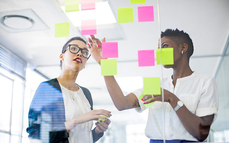 Zwei Geschäftsfrauen stehen vor einer Glaswand mit Haftnotizen und sprechen über Zeitmanagement.