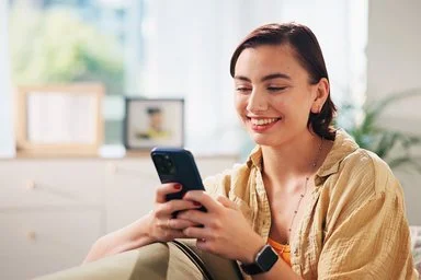 Eine lächelnde junge Frau sitzt auf einem Sofa und hält ein Smartphone in beiden Händen.