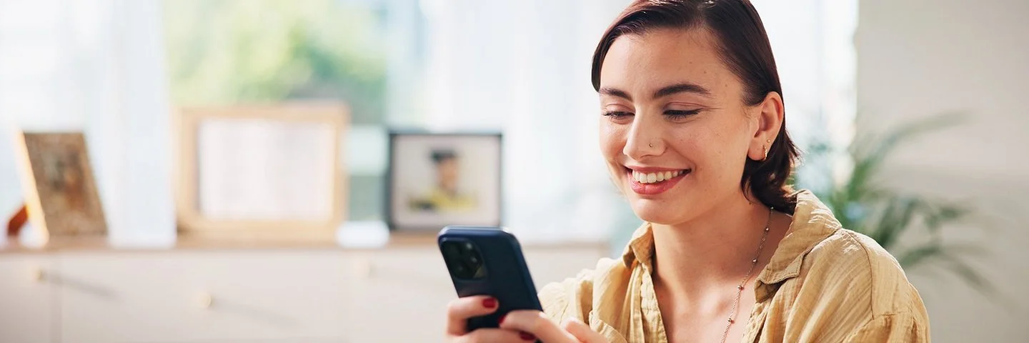 Eine lächelnde junge Frau sitzt auf einem Sofa und hält ein Smartphone in beiden Händen.
