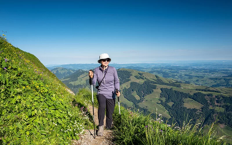 Eine Frau besteigt bei schönem Sommerwetter mit Wanderstöcken einen Berg. Sie trägt dabei Sonnenbrille, Sonnenhut und lange dünne Kleidung.