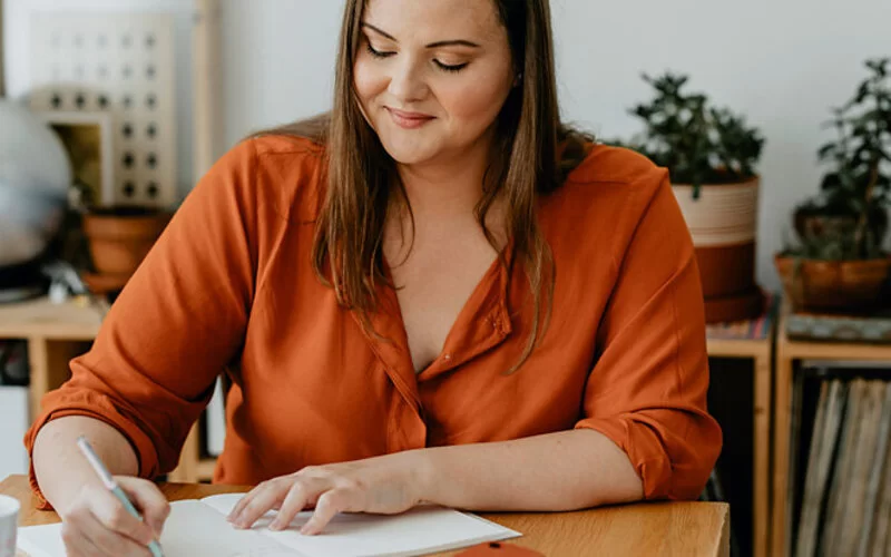 Eine Frau sitzt an einem Schreibtisch und hält in der rechten Hand einen Stift, mit dem sie etwas in ein Buch notiert.