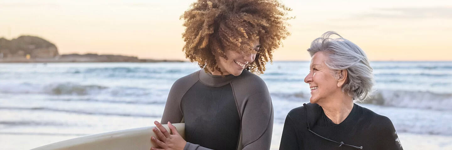 Eine junge und eine ältere Frau lachen sich am Strand an, unter ihren Armen tragen sie ein Surfboard.