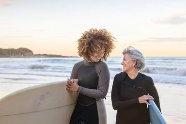 Eine junge und eine ältere Frau lachen sich am Strand an, unter ihren Armen tragen sie ein Surfboard.
