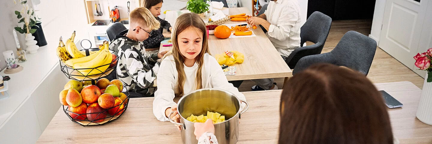Eine Familienküche. Im Vordergrund ein ungefähr zehnjähriges Mädchen, das einen großen Topf mit geschnittenen Kartoffeln in den Händen hält. Hinter ihr am Tisch sitzen ein Junge und ein Mädchen im Teenageralter sowie ein Mann mit Bart, sie schneiden Karotten und Kürbis. 
