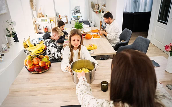 Eine Familienküche. Im Vordergrund ein ungefähr zehnjähriges Mädchen, das einen großen Topf mit geschnittenen Kartoffeln in den Händen hält. Hinter ihr am Tisch sitzen ein Junge und ein Mädchen im Teenageralter sowie ein Mann mit Bart, sie schneiden Karotten und Kürbis. 