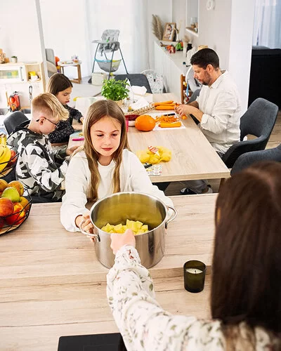 Eine Familienküche. Im Vordergrund ein ungefähr zehnjähriges Mädchen, das einen großen Topf mit geschnittenen Kartoffeln in den Händen hält. Hinter ihr am Tisch sitzen ein Junge und ein Mädchen im Teenageralter sowie ein Mann mit Bart, sie schneiden Karotten und Kürbis. 
