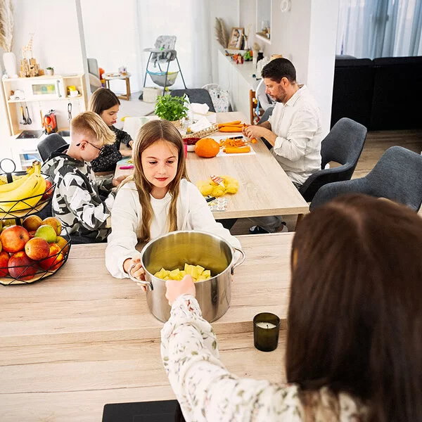 Eine Familienküche. Im Vordergrund ein ungefähr zehnjähriges Mädchen, das einen großen Topf mit geschnittenen Kartoffeln in den Händen hält. Hinter ihr am Tisch sitzen ein Junge und ein Mädchen im Teenageralter sowie ein Mann mit Bart, sie schneiden Karotten und Kürbis.