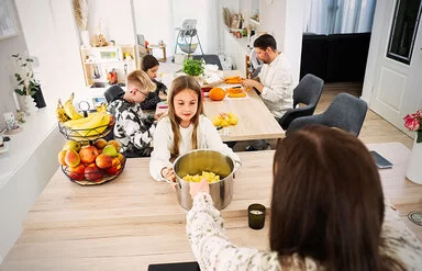 Eine Familienküche. Im Vordergrund ein ungefähr zehnjähriges Mädchen, das einen großen Topf mit geschnittenen Kartoffeln in den Händen hält. Hinter ihr am Tisch sitzen ein Junge und ein Mädchen im Teenageralter sowie ein Mann mit Bart, sie schneiden Karotten und Kürbis.