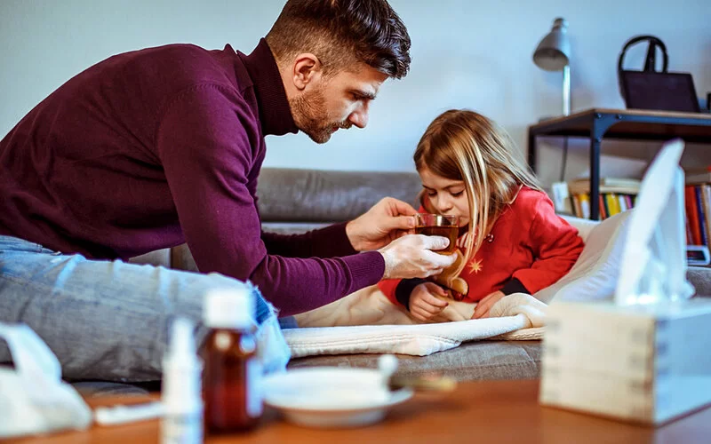 Ein kleines erkältetes Mädchen liegt auf der Couch und bekommt von ihrem Vater eine Tasse Tee zum Trinken gereicht.