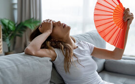 Eine Frau leidet unter Kreislaufproblemen durch Hitze und sitzt erschöpft auf einem Sofa, während sie sich Luft mit einem Fächer zu wedelt.