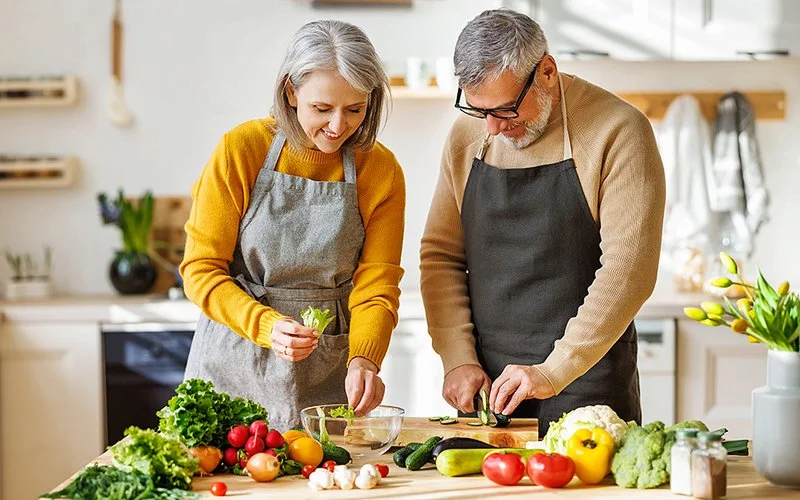 Ein Mann und eine Frau tragen Kochschürzen und stehen in einer Küche. Sie schneiden Gemüse für eine gesunde Mahlzeit.
