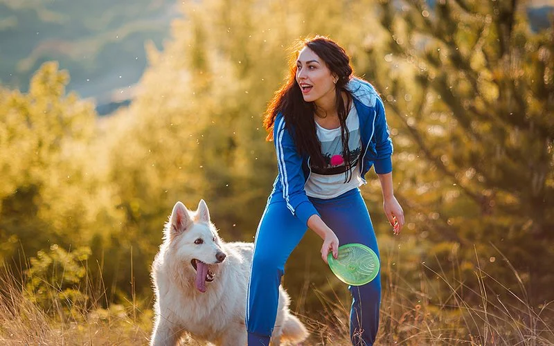 Eine Frau in einem blauen Jogginganzug wirft eine grüne Frisbee, daneben steht ein Hund.