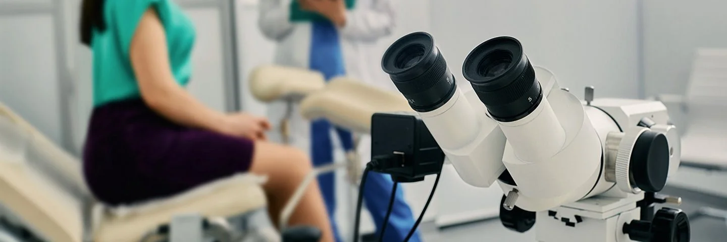 Eine Frau in einer gynäkologischen Praxis im Gespräch mit ihrer Ärztin, im Bildvordergrund ein Kolposkop.