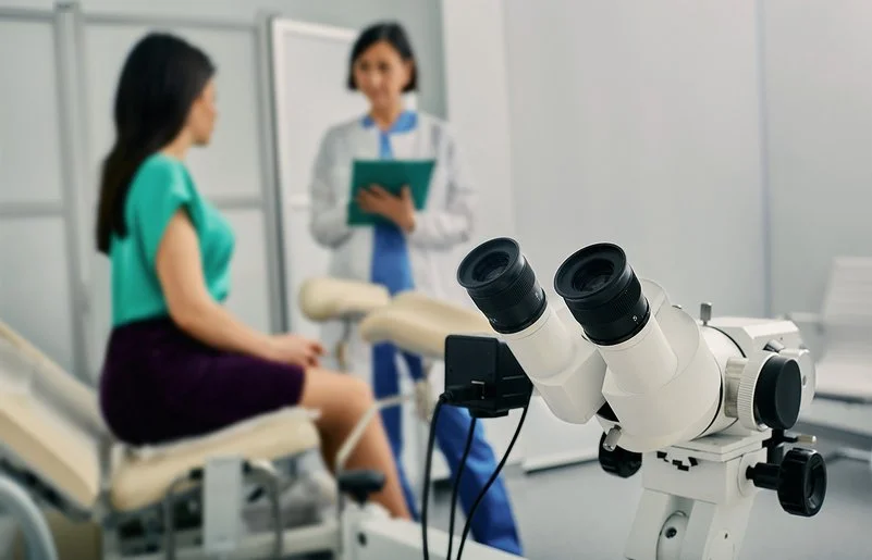 Eine Frau in einer gynäkologischen Praxis im Gespräch mit ihrer Ärztin, im Bildvordergrund ein Kolposkop.