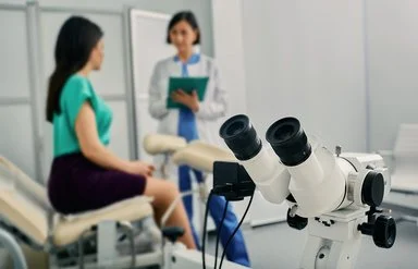 Eine Frau in einer gynäkologischen Praxis im Gespräch mit ihrer Ärztin, im Bildvordergrund ein Kolposkop.