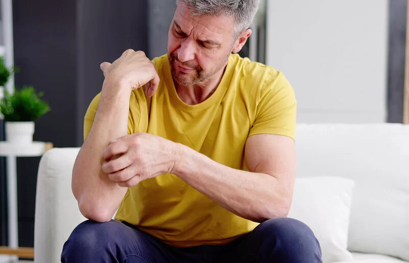 Ein Mann mit gelbem T-Shirt sitzt auf einem Sofa und kratzt sich an seinem Unterarm.
