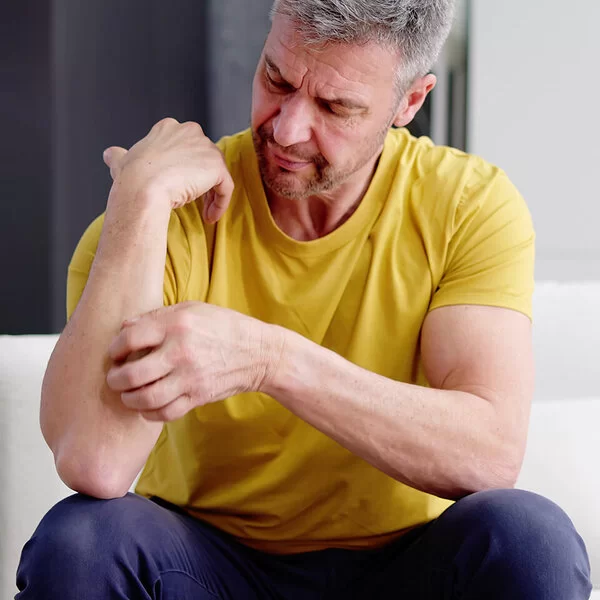 Ein Mann mit gelbem T-Shirt sitzt auf einem Sofa und kratzt sich an seinem Unterarm.