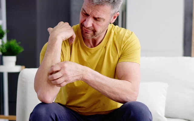 Ein Mann mit gelbem T-Shirt sitzt auf einem Sofa und kratzt sich an seinem Unterarm.