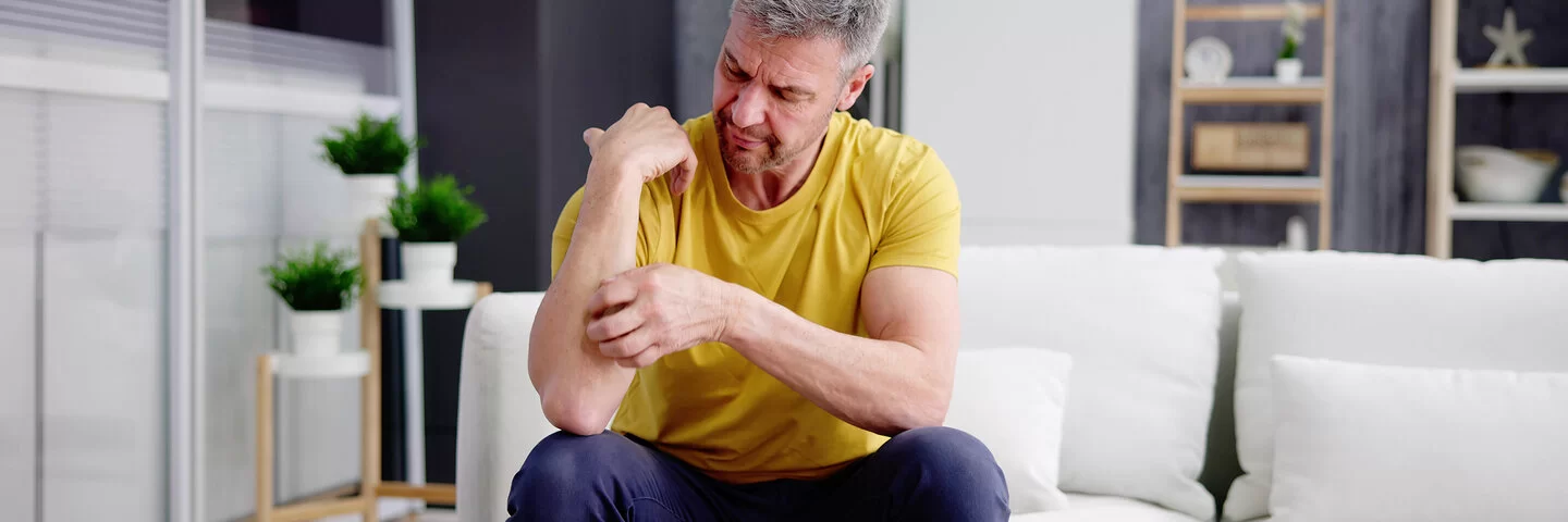 Ein Mann mit gelbem T-Shirt sitzt auf einem Sofa und kratzt sich an seinem Unterarm.