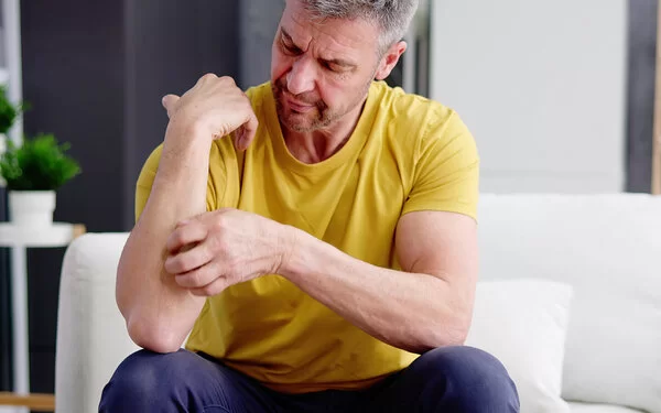 Ein Mann mit gelbem T-Shirt sitzt auf einem Sofa und kratzt sich an seinem Unterarm.