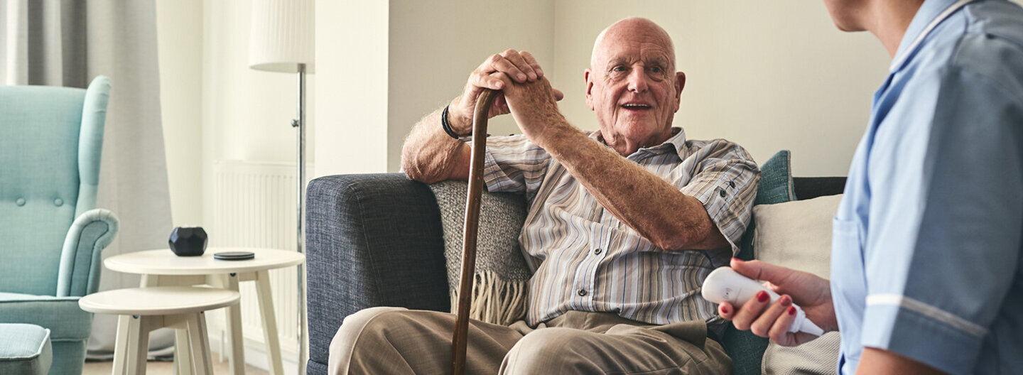 Pflege im Alltag: Ein älterer Mann sitzt lächelnd auf einem Sofa und unterhält sich mit einer weiblichen Betreuungsperson.