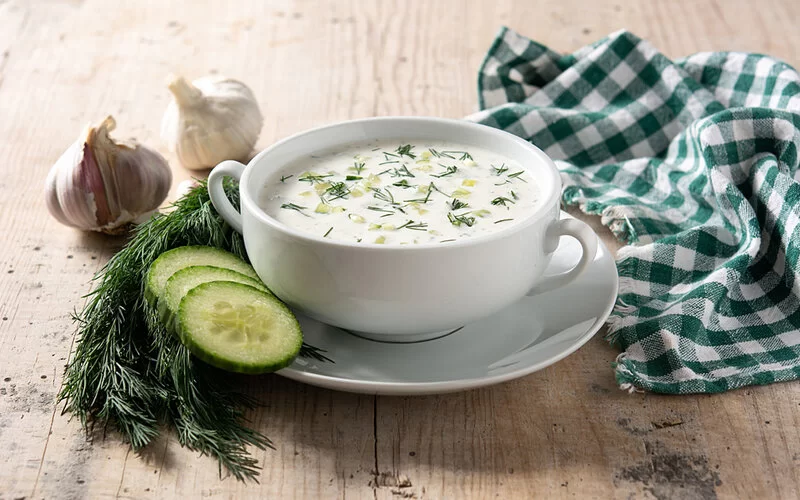 Ein Tässchen Gurken-Joghurt-Suppe mit Gurkenscheiben auf der Untertasse steht auf einem Holztisch.