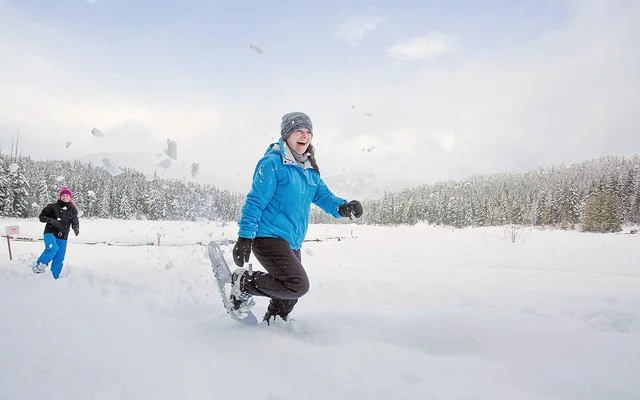 Zwei Frauen wandern lächelnd mit Schneeschuhen durch eine winterliche Landschaft.