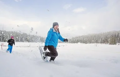 Zwei Frauen wandern lächelnd mit Schneeschuhen durch eine winterliche Landschaft.