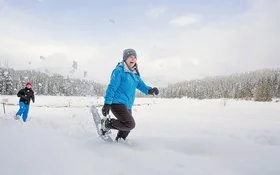 Zwei Frauen wandern lächelnd mit Schneeschuhen durch eine winterliche Landschaft.