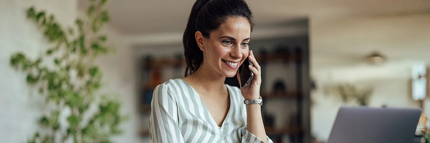 Eine Frau mit zusammengebundenen dunklen Haaren sitzt an einem Holztisch und telefoniert mit einem Smartphone. Sie trägt eine hell gestreifte Bluse mit V-Ausschnitt. Vor ihr steht ein aufgeklappter Laptop, den sie mit einer Hand bedient. Im Hintergrund sind eine große grüne Zimmerpflanze und unscharfe Einrichtungsgegenstände zu erkennen. Die Szene wirkt hell und modern.