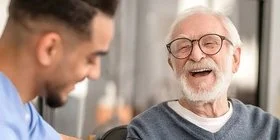 Ein älterer Mann sitzt in einem Wohnzimmer auf einem Stuhl. Neben ihm sitzt ein Pfleger, der dem älteren Mann mit einem Igel-Ball die rechte Handinnenfläche mobilisiert. Beide lachen dabei.