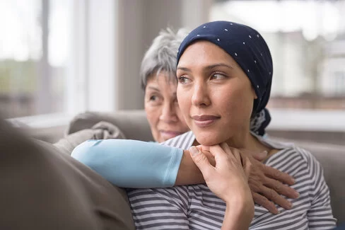 Eine jüngere Krebs-Patientin und eine ältere Frau sitzen innig auf dem Sofa.
