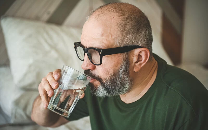 Ein älterer Mann mit Brille und grauem Bart sitzt auf dem Bett und trinkt Wasser aus einem Glas.